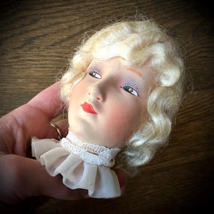 May include: A vintage doll head with a painted face, red lips, and blonde curly hair. The doll head has a white lace collar and is set against a dark wooden background. The doll's eyes are painted with long eyelashes.