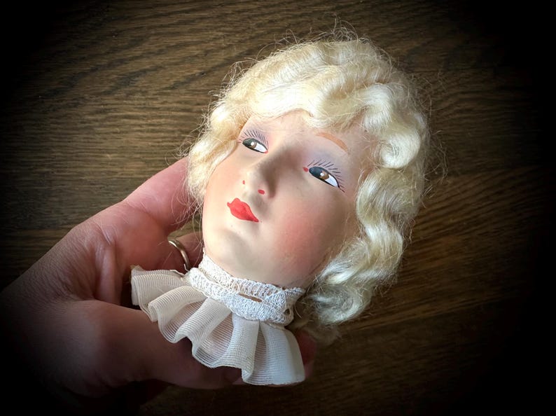 May include: A vintage doll head with a painted face, red lips, and blonde curly hair. The doll head has a white lace collar and is set against a dark wooden background. The doll's eyes are painted with long eyelashes.