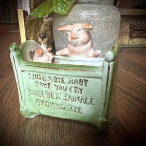 May include: A green ceramic planter shaped like a crib with the text "Hush-a-bye baby don't you cry you'll be a sausage by and by". Two small pig figurines are inside the planter.