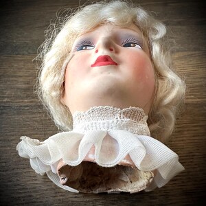 May include: A vintage doll head with blonde, curly hair, painted blue eyes, and red lips. The doll head has a white lace collar and a pink fabric base. The doll head is on a wooden surface.