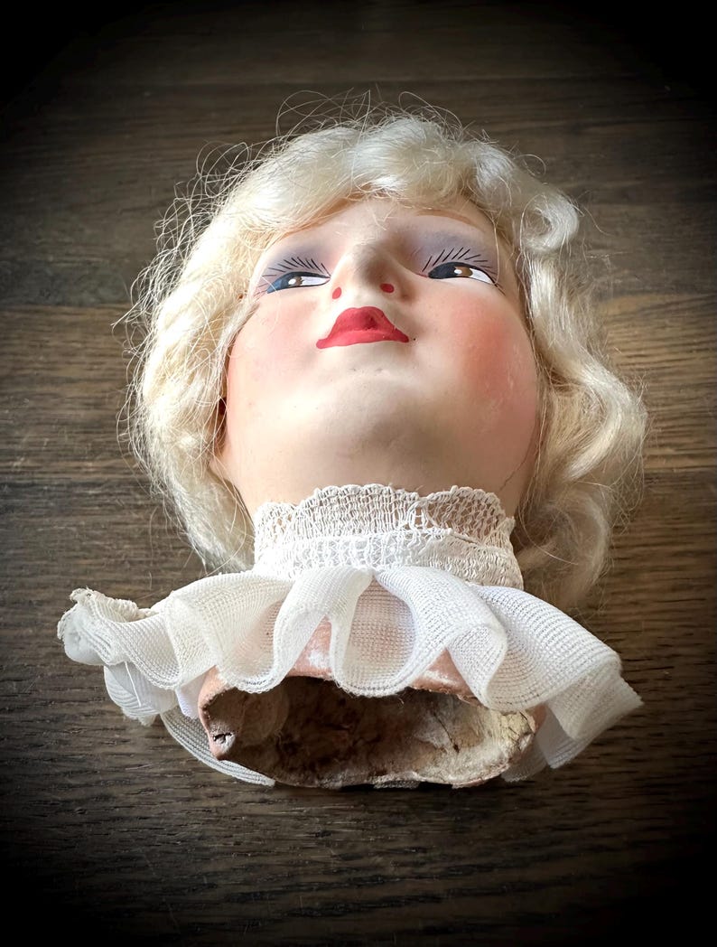 May include: A vintage doll head with blonde, curly hair, painted blue eyes, and red lips. The doll head has a white lace collar and a pink fabric base. The doll head is on a wooden surface.