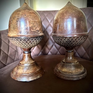 May include: Two antique-style acorn-shaped decorative objects. The tops are smooth and brown, while the bases feature a detailed diamond pattern. The objects sit on a pedestal base, and are made of a dark, aged material.