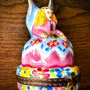 Puede incluir: Caja de baratijas de cerámica pintada a mano con una figura sentada con cabello rubio y un vestido rosa. La caja tiene un diseño floral con un borde dorado y un cierre en forma de estrella. Fondo de madera.