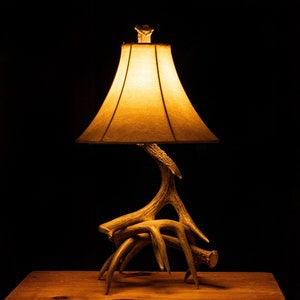 May include: A table lamp with a beige lampshade and a base made of antlers. The lamp is turned on and casting a warm glow.
