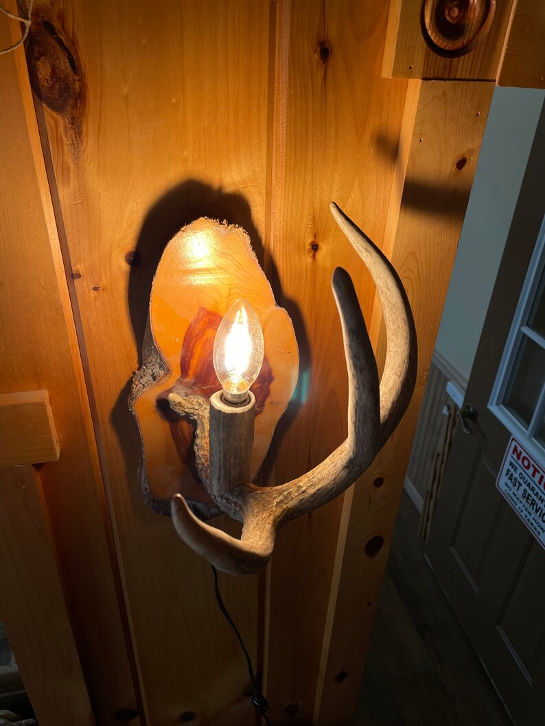 Op de afbeelding: Een houten wandlamp met een gloeilamp en hertengewei accenten. De lamp is gemonteerd op een houten muur.