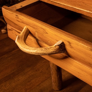 May include: A wooden drawer with a natural antler handle. The drawer is made of light brown wood with visible grain, and the handle is a curved piece of antler, adding a rustic touch. The drawer is open, revealing a dark interior.