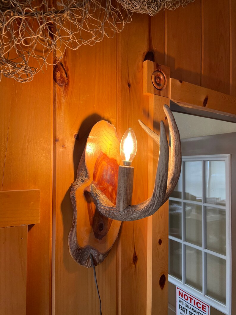 Op de afbeelding: Een houten wandlamp met een gloeilamp en een hertengewei-ontwerp. De lamp is gemonteerd op een houten muur en wordt verlicht door een gloeilamp. De lamp is gemaakt van natuurlijke materialen en heeft een rustieke uitstraling.