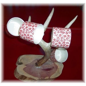 May include: A set of four white ceramic mugs with a red floral pattern, displayed on a wooden base with antler handles.