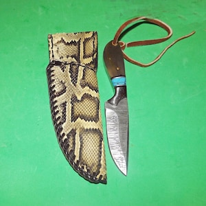 May include: A handmade knife with a patterned blade and a handle with turquoise accents. The knife has a brown leather strap and is next to a snakeskin sheath. The sheath is black, tan, and brown.