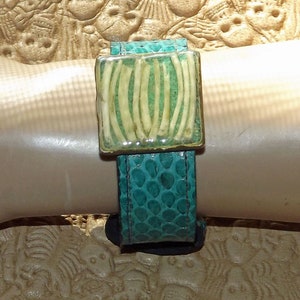 May include: A turquoise blue leather bracelet with a clear resin square charm featuring a white bone pattern.