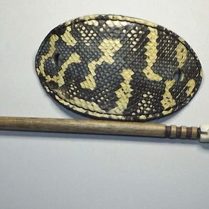 May include: A brown and black oval-shaped hair stick with a decorative, multi-colored, bone-like tip. The stick is lying on a white surface next to a brown and yellow oval-shaped hair clip.