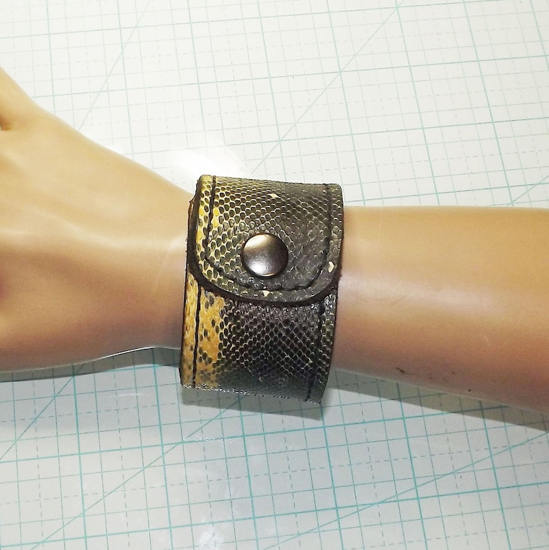 May include: A brown leather cuff bracelet with a snakeskin pattern and a silver button closure.