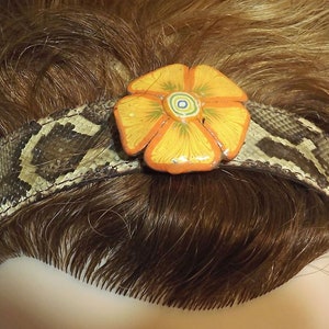 May include: A brown snakeskin headband with a yellow flower accent. The flower has orange petals and a multi-colored center.