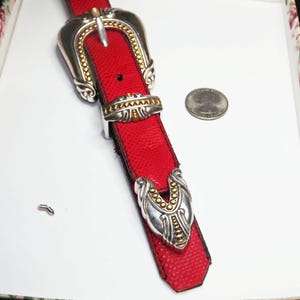 May include: A red leather belt with a silver and gold buckle and accents. The belt has a textured pattern and a decorative silver and gold clasp. A quarter coin is next to the belt.