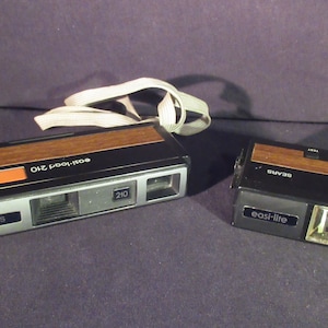 May include: A vintage Sears Easl-Lite 210 camera with a brown woodgrain finish and a black body. The camera has a built-in flash and a strap.