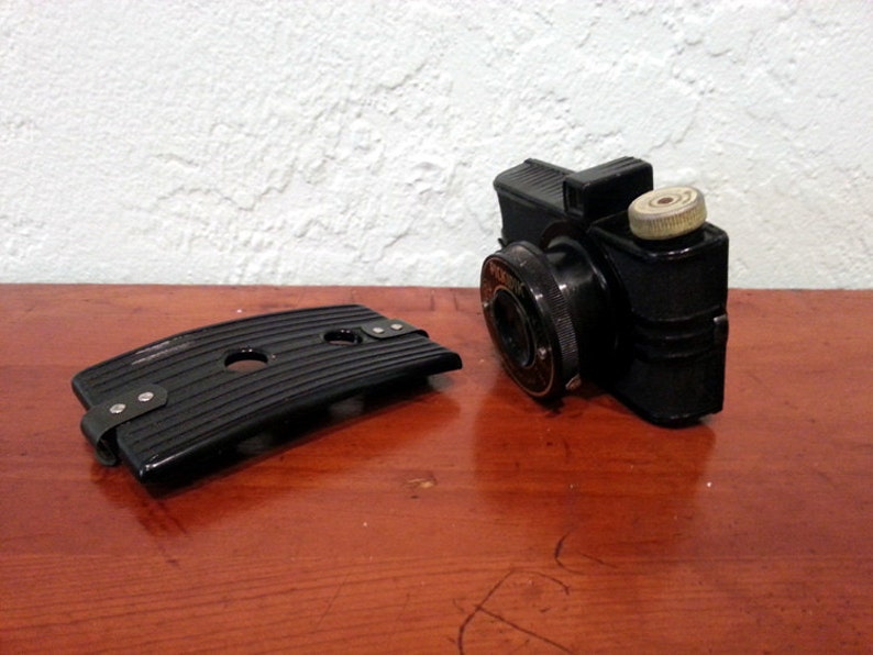 Peut inclure: Un appareil photo vintage en plastique noir avec une plaque arri&egrave;re amovible. L'appareil photo a un objectif rond et un petit bouton rond blanc cass&eacute; sur le dessus.