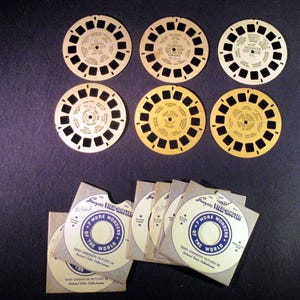 May include: A collection of vintage View-Master reels and their corresponding paper sleeves. The reels are circular with multiple small viewing windows and text. The sleeves are white with blue text and graphics, and the text reads "7 More Wonders of the World."