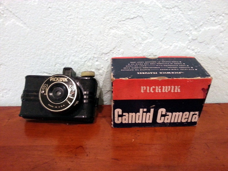 Peut inclure: Un appareil photo Pickwick Candid Camera noir avec un objectif dor&eacute; et les mots "Pickwick Candid Camera" sur la bo&icirc;te.