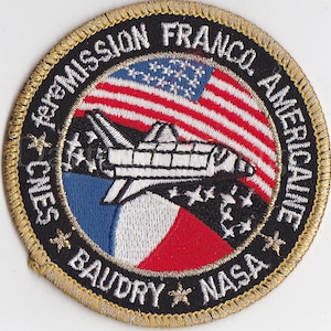 May include: A black and gold embroidered patch with the text "Mission Franco-Americaine" and "CNES BAUDRY NASA" surrounding a white space shuttle flying over a red, white, and blue American flag.