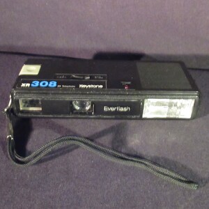 May include: A black Keystone XR 308 2x Telephoto camera with a built-in Everflash. The camera has a black strap attached.