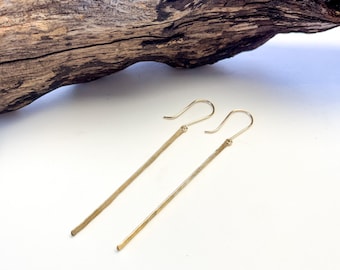 May include: A pair of long, thin gold earrings with a hammered finish. The earrings are hanging from a piece of driftwood.