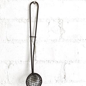 May include: A dark brown metal skimmer with a long, slender handle and a circular, wire-mesh bowl. The tool features a simple, utilitarian design with a loop at the top of the handle for hanging, constructed from thin, dark-toned metal wire.