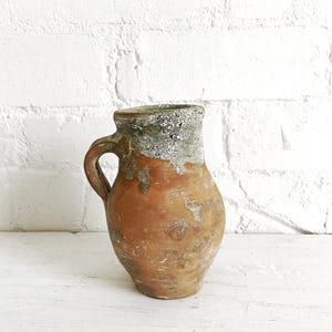 May include: A rustic terracotta pitcher featuring a bulbous body and a single curved handle. The upper portion of the vessel is coated in a textured, weathered greyish-green glaze that drips down onto the natural, unglazed reddish-brown clay surface.