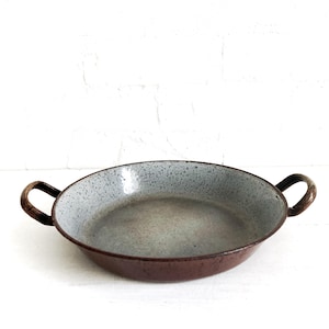 May include: A round, shallow metal pan with two curved side handles. The interior features a light grey speckled finish, while the exterior and handles are a solid dark brown colour. The pan has a wide, open design with a flat base.