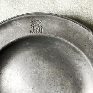 May include: A close-up of a pewter plate with a decorative emblem. The plate is a muted gray color with a slightly reflective surface. The emblem appears to be a monogram with a crown above it. The plate has a simple, classic design.