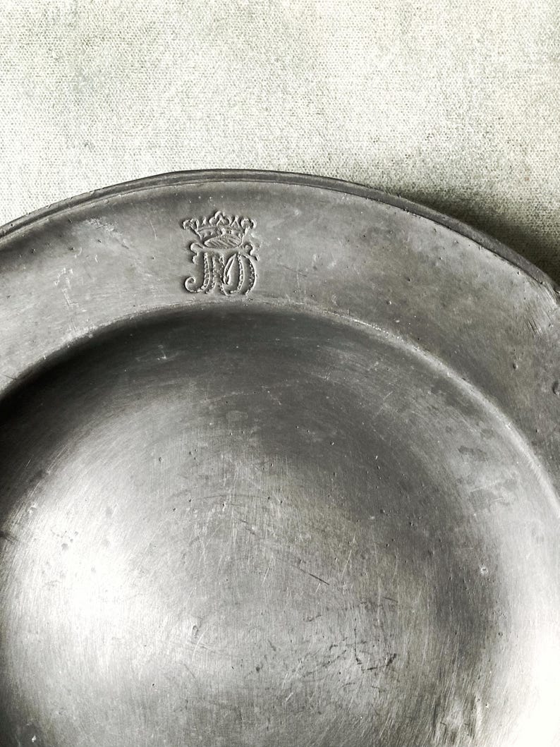 May include: A close-up of a pewter plate with a decorative emblem. The plate is a muted gray color with a slightly reflective surface. The emblem appears to be a monogram with a crown above it. The plate has a simple, classic design.
