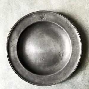 May include: A weathered, round, pewter plate with a wide rim. The plate has a slightly concave center and a small, embossed emblem near the top. The plate's surface shows signs of age and use, with subtle variations in tone.
