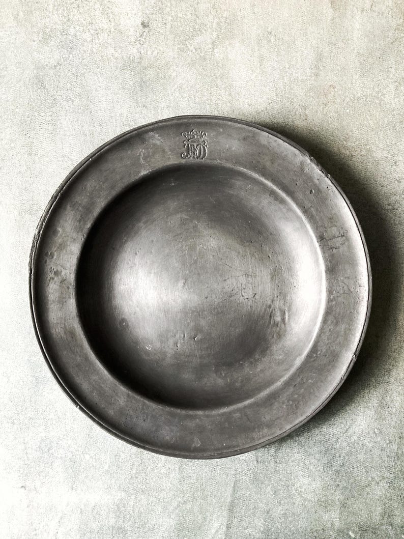May include: A weathered, round, pewter plate with a wide rim. The plate has a slightly concave center and a small, embossed emblem near the top. The plate's surface shows signs of age and use, with subtle variations in tone.