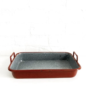 May include: A rectangular, vintage-style baking pan with a red enamel exterior and two small handles. The interior has a textured, grey surface. The pan is set against a white brick wall.