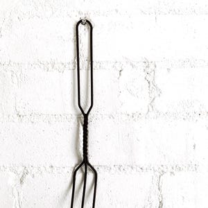 May include: A dark brown, hand-forged metal fork featuring a long, slender handle with a looped top for hanging. The central section of the handle displays a decorative twisted metal design, leading down to three sharp, pointed tines at the base.