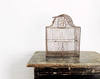 Op de afbeelding: Een antieke, decoratieve vogelkooi gemaakt van dun, verouderd metaaldraad. De kooi heeft een gebogen bovenkant met een bloemenontwerp en staat op een verweerde, donkere houten tafel. De achtergrond is een witte muur met horizontale panelen.