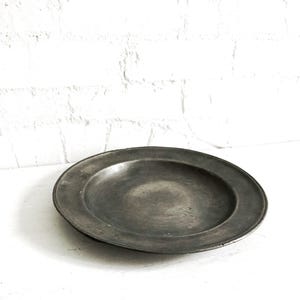 May include: A round, antique pewter plate with a wide rim. The plate has a dark, matte finish and a slightly concave center. The background is a white surface and a white brick wall.