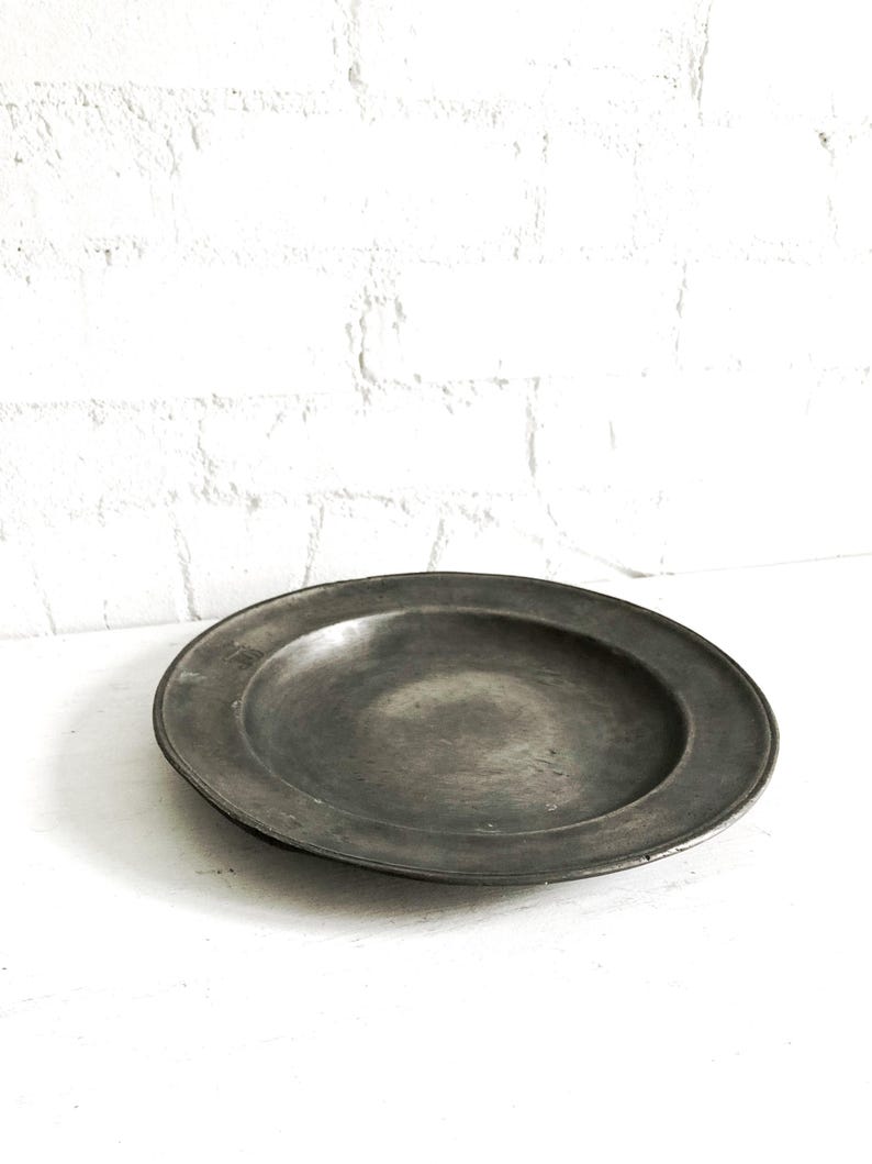 May include: A round, antique pewter plate with a wide rim. The plate has a dark, matte finish and a slightly concave center. The background is a white surface and a white brick wall.