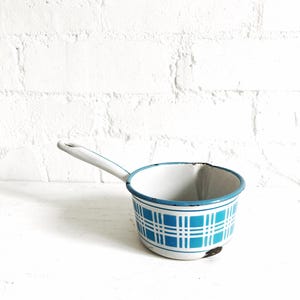 May include: A small, vintage enamel saucepan with a white handle and a blue and white plaid pattern. The rim and handle are trimmed in blue. The saucepan is set against a white background.