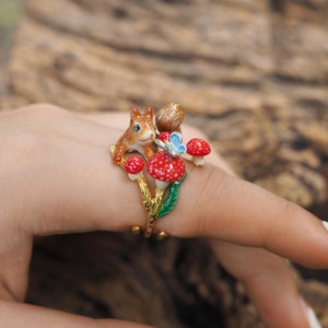 May include: A gold ring with a squirrel, mushrooms, and a butterfly. The squirrel is brown and has a white belly. The mushrooms are red with white spots. The butterfly is blue with white spots.