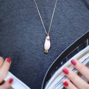 May include: A woman wearing a gray shirt with a gold chain necklace featuring a pink bird pendant. She is holding a stack of magazines.