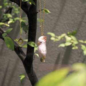 May include: A gold chain necklace with a pink and white porcelain cockatoo pendant hanging from a branch.