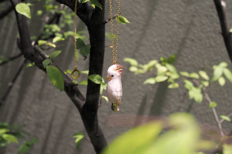 May include: A gold chain necklace with a pink and white porcelain cockatoo pendant hanging from a branch.