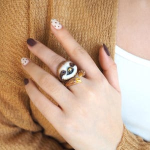 May include: A ring featuring a sculpted sloth with brown fur, a white face, and dark eye patches. The sloth is depicted in a climbing position with its front paws wrapped around the band, showcasing detailed golden claws and a smiling expression.