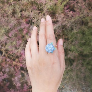 May include: A ring featuring a blue flower with green leaves. The ring is on a finger against a blurred background of pink and green foliage.