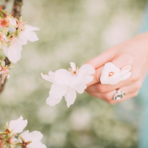 May include: A white ceramic bunny rabbit figurine with a red dot on its nose. The figurine is being held by a hand with a silver ring.