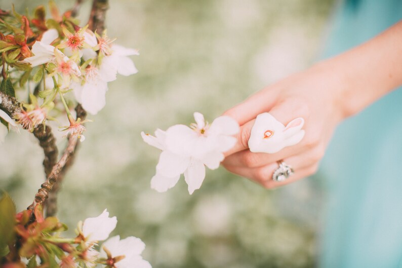May include: A white ceramic bunny rabbit figurine with a red dot on its nose. The figurine is being held by a hand with a silver ring.
