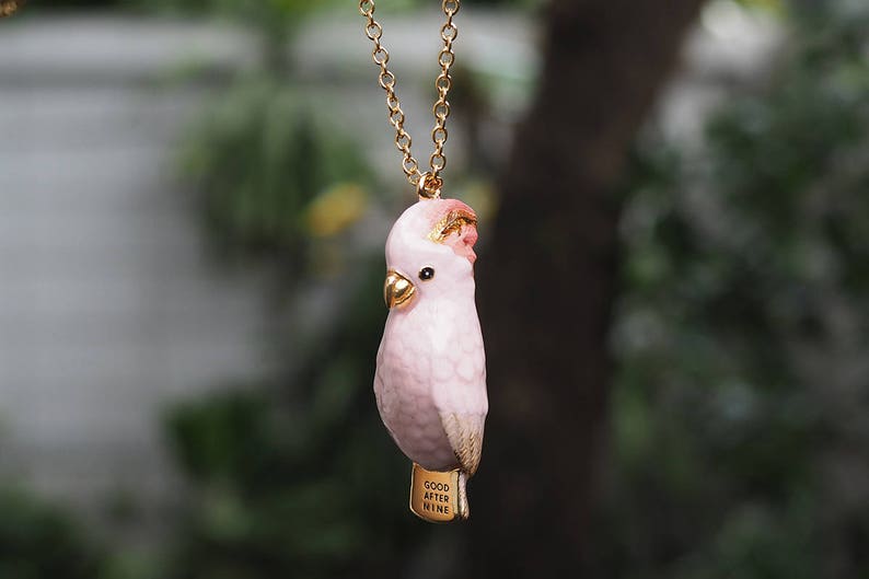 May include: A pink cockatoo bird charm necklace with a gold chain and the words "Good After Nine" on the charm.