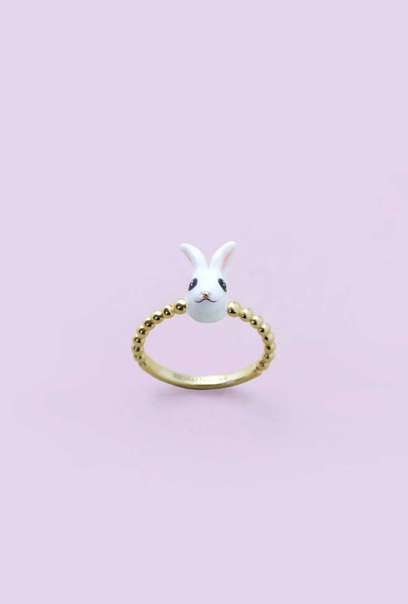 K&ouml;nnte beinhalten: Ein goldfarbener Ring mit einem wei&szlig;en Hasenkopf mit schwarzen Augen und rosa Ohren. Das Ringband ist mit kleinen, goldfarbenen Perlen verziert. Der Hintergrund ist helllila.