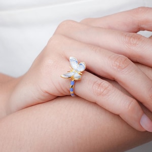 Könnte beinhalten: Ein goldener Ring mit einem blauen und weißen Schmetterlingsdesign. Der Schmetterling ist mit weißem Emaille verziert und hat einen goldenen Umriss. Die Ringschiene ist blau und gold.