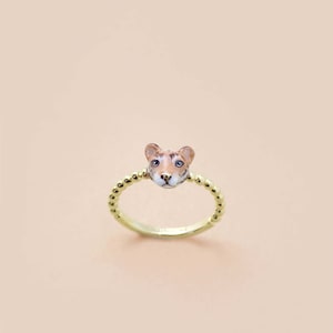 Puede incluir: Un anillo de oro con una banda de cuentas y un dije de cabeza de tigre. El tigre tiene pelaje marrón y naranja y ojos azules.